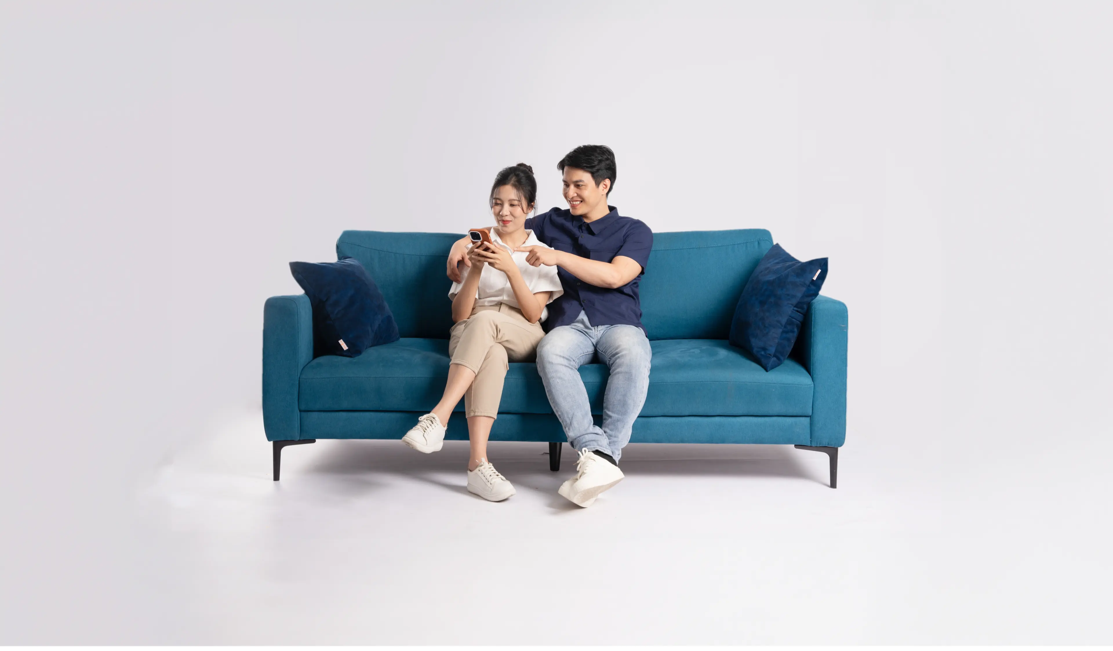 A young man and a young woman sitting on a couch happily looking at a cellphone which the latter is holding