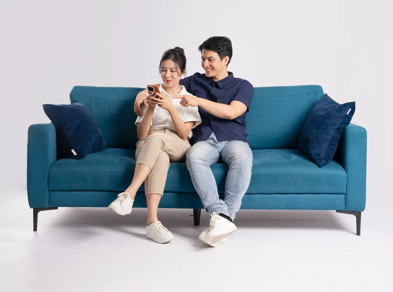 A young man and a young woman sitting on a couch happily looking at a cellphone which the latter is holding